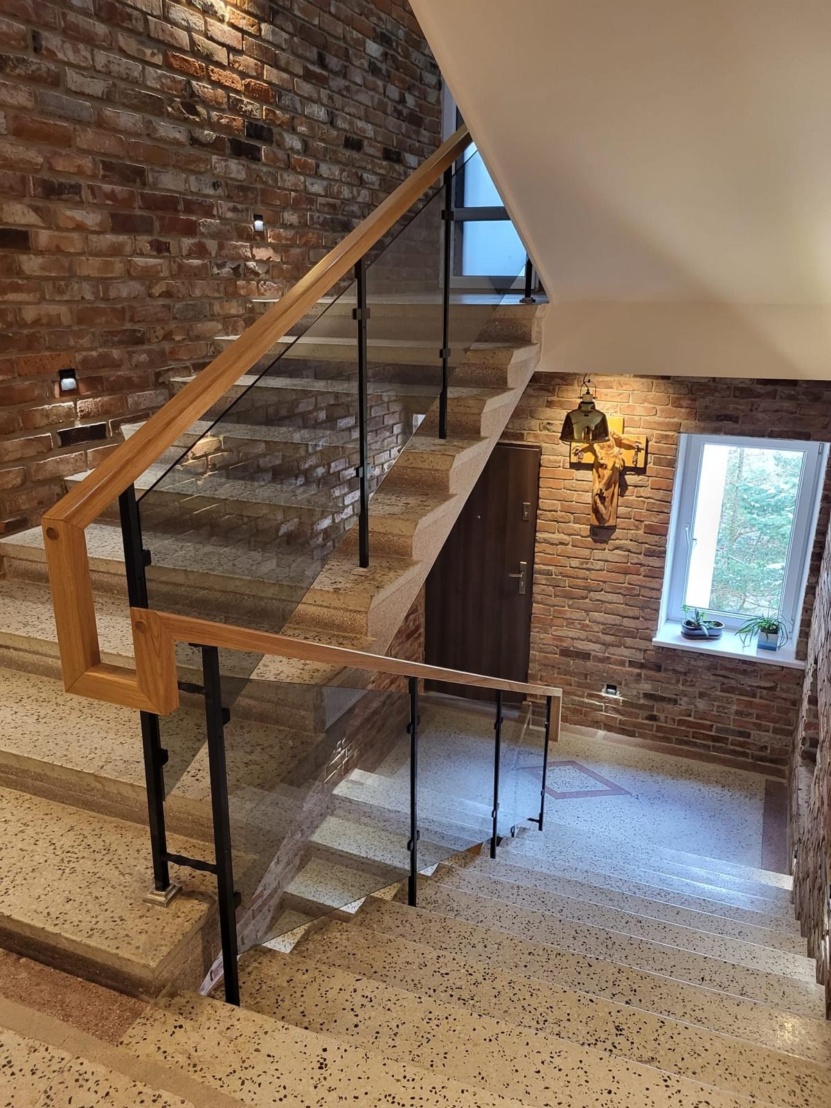Szklane balustrady schodowe z drewnianymi poręczami i czarnymi wspornikami w budynku z ceglanymi ścianami. Widok z góry na dół.