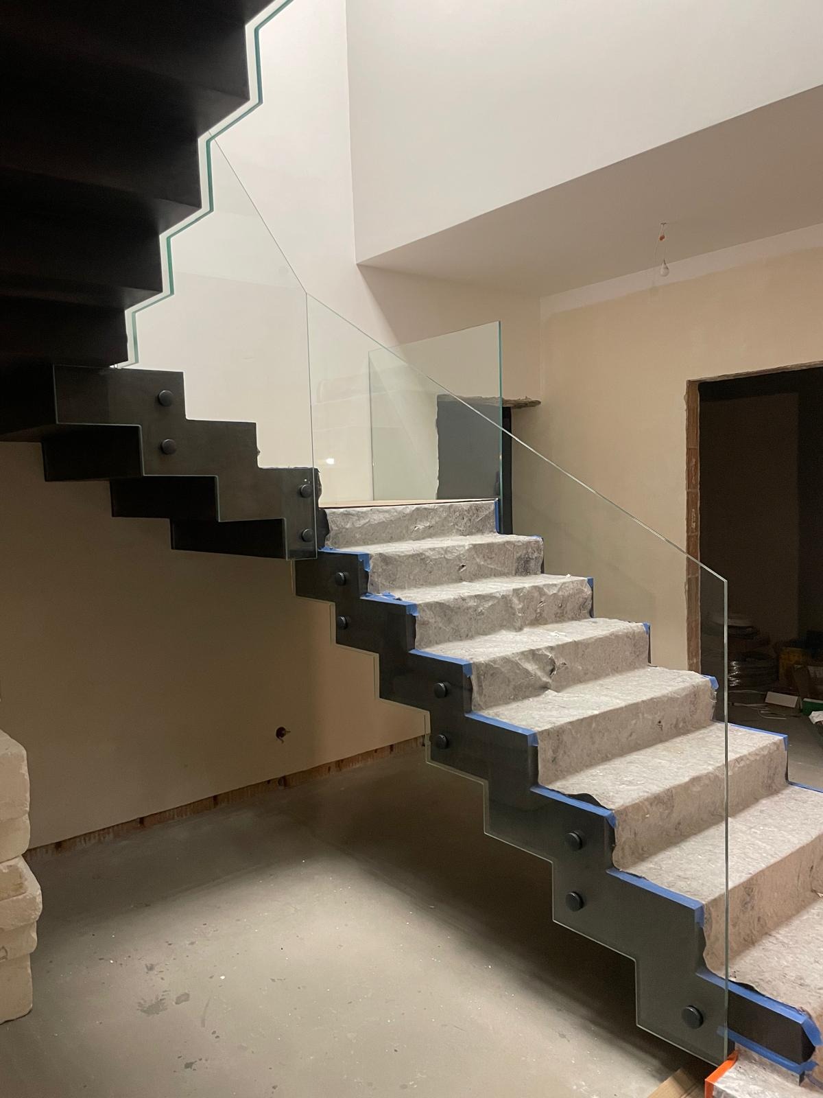 Szklana balustrada na schodach z szarego kamienia, zamontowana na czarnych wspornikach, zabezpieczona folią, podczas remontu wnętrza.