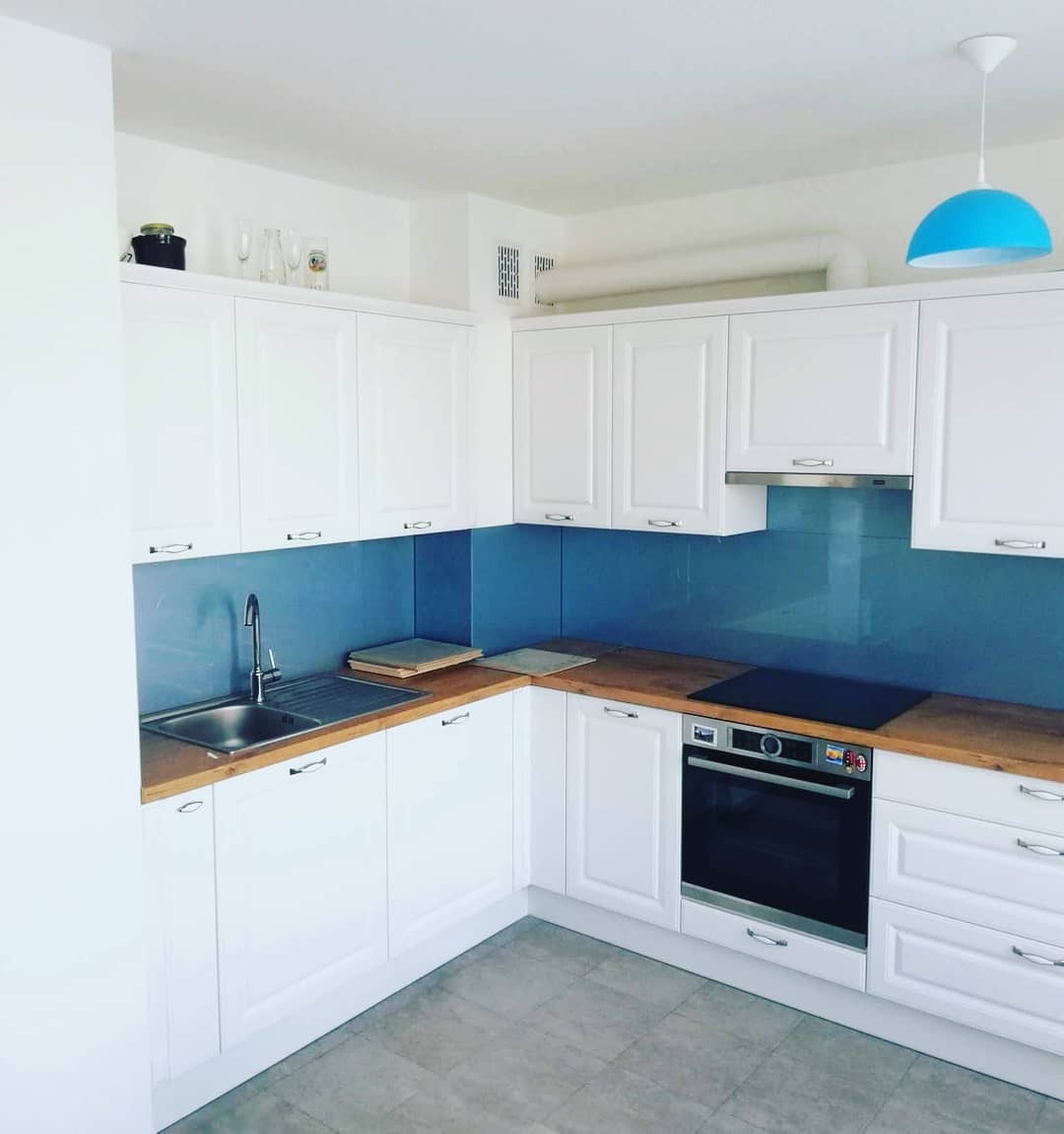 Błękitny panel szklany nad blatem w białej kuchni. Nowoczesny design z wiszącą lampą i drewnianym blatem. Fronty szafek frezowane.