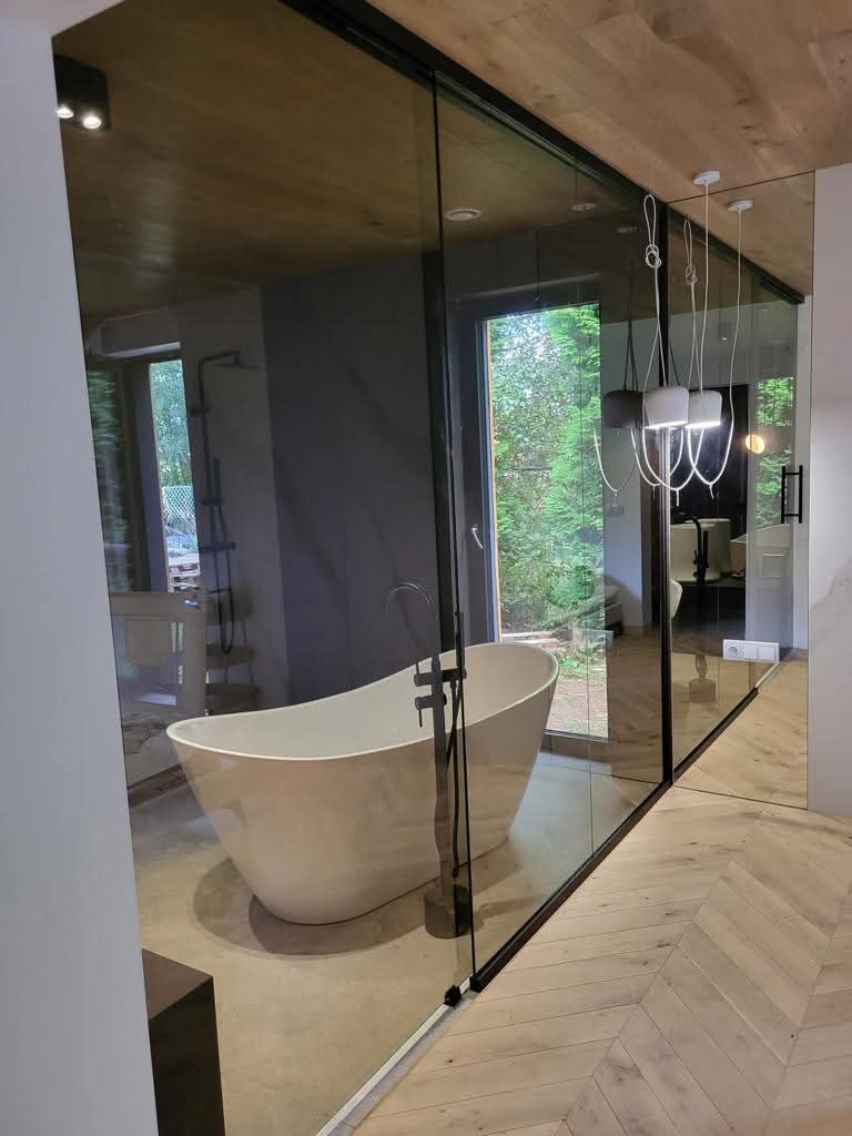 Szklana ściana działowa w łazience z wolnostojącą wanną. Widoczne detale wykończenia, drewniany sufit i parkiet ułożony w jodełkę. Nowoczesny design.
