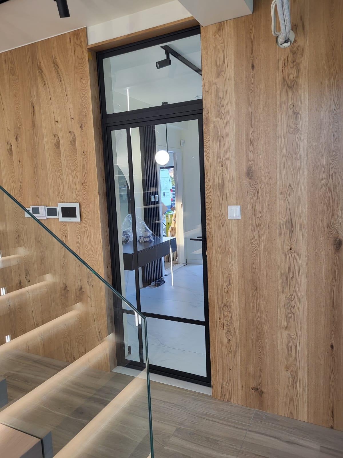 Nowoczesne, przeszklone drzwi z czarną ramą, widoczne z perspektywy schodów z podświetleniem LED. Ściany wykończone drewnopodobnymi panelami.