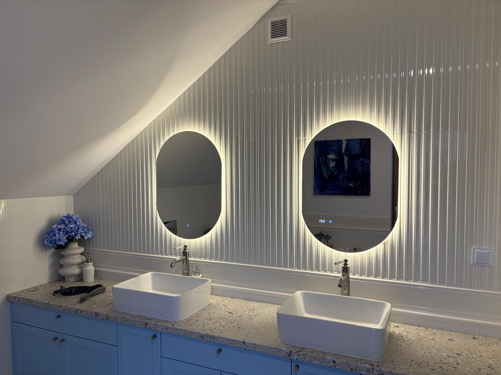 Nowoczesna łazienka na poddaszu z dwoma owalnymi lustrami LED, białymi umywalkami i niebieskimi szafkami. Ściany wyłożone białymi panelami.