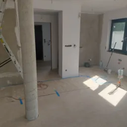 Wnętrze w trakcie remontu: kolumna, drabina, odsłonięte instalacje elektryczne, przygotowane ściany do malowania, zabezpieczona podłoga. Widoczne narzędzia i materiały budowlane.