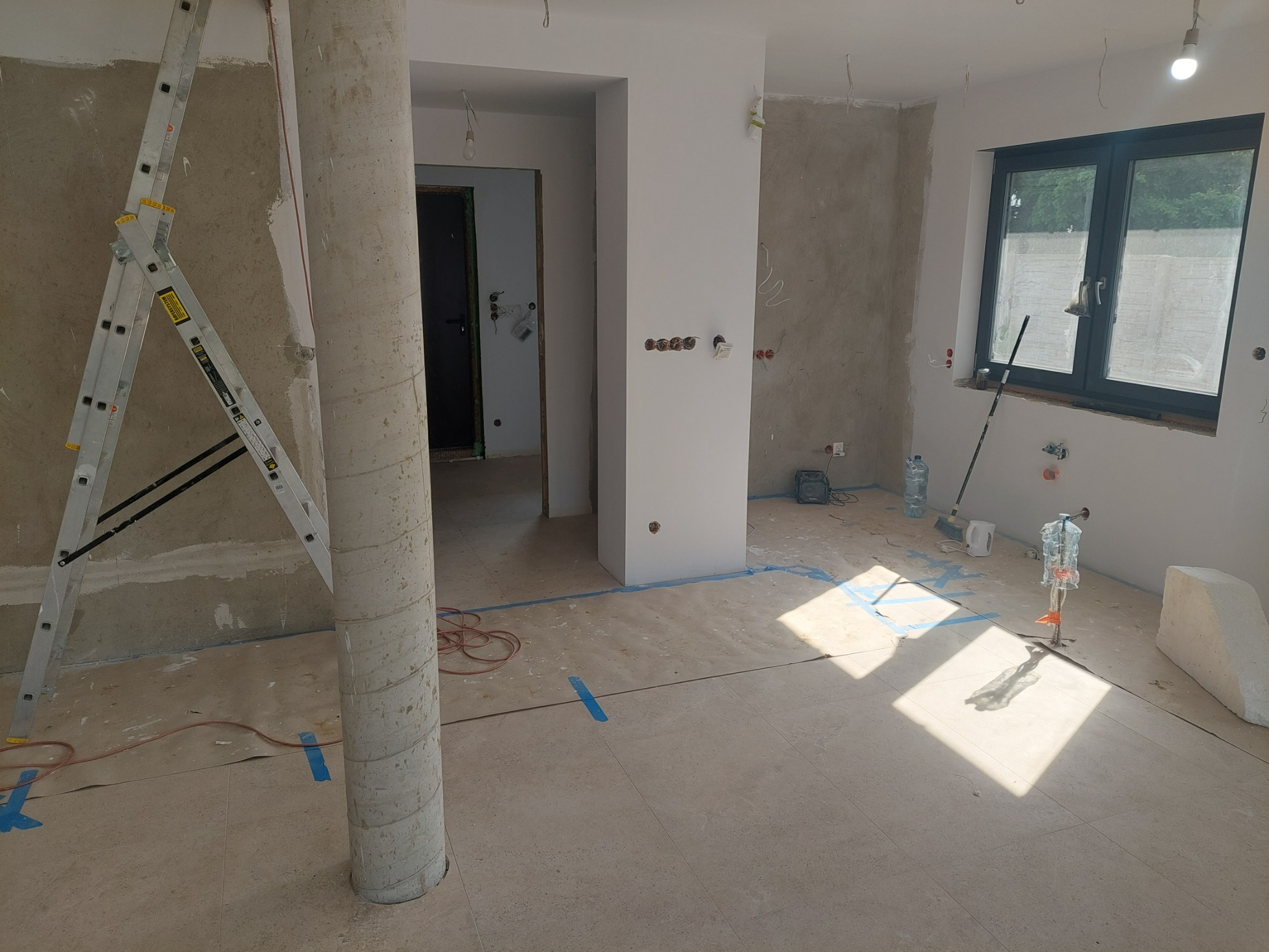Wnętrze w trakcie remontu: kolumna, drabina, odsłonięte instalacje elektryczne, przygotowane ściany do malowania, zabezpieczona podłoga. Widoczne narzędzia i materiały budowlane.
