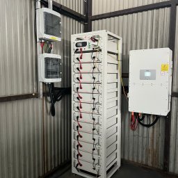 INTELUS GROUP Sp. z o.o. - Biała szafa z akumulatorami magazynu energii, skrzynki elektryczne i inwerter na ścianie z blachy falistej w pomieszczeniu. Instalacja gotowa do użytku.