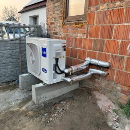 INTELUS GROUP Sp. z o.o. - Biała jednostka zewnętrzna klimatyzacji Haier, zamontowana na betonowych podstawach przy ceglanej ścianie budynku. Widoczne ocieplone rury instalacji. Na dachu panele fotowoltaiczne.