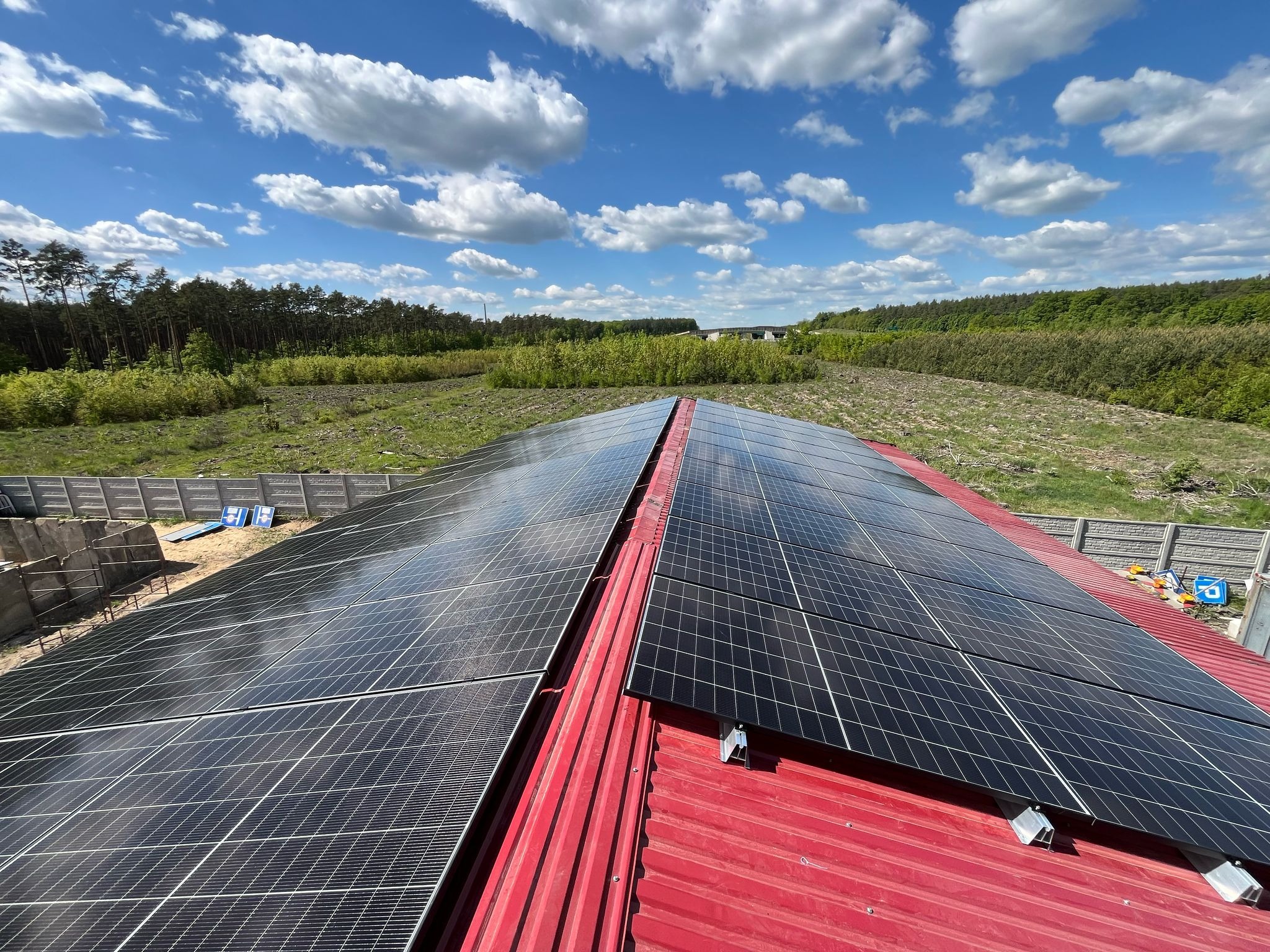 Instalacja paneli fotowoltaicznych na czerwonym dachu z blachy trapezowej. W tle błękitne niebo z chmurami i zieleń drzew. Widok z góry, ukazujący gotowy system.
