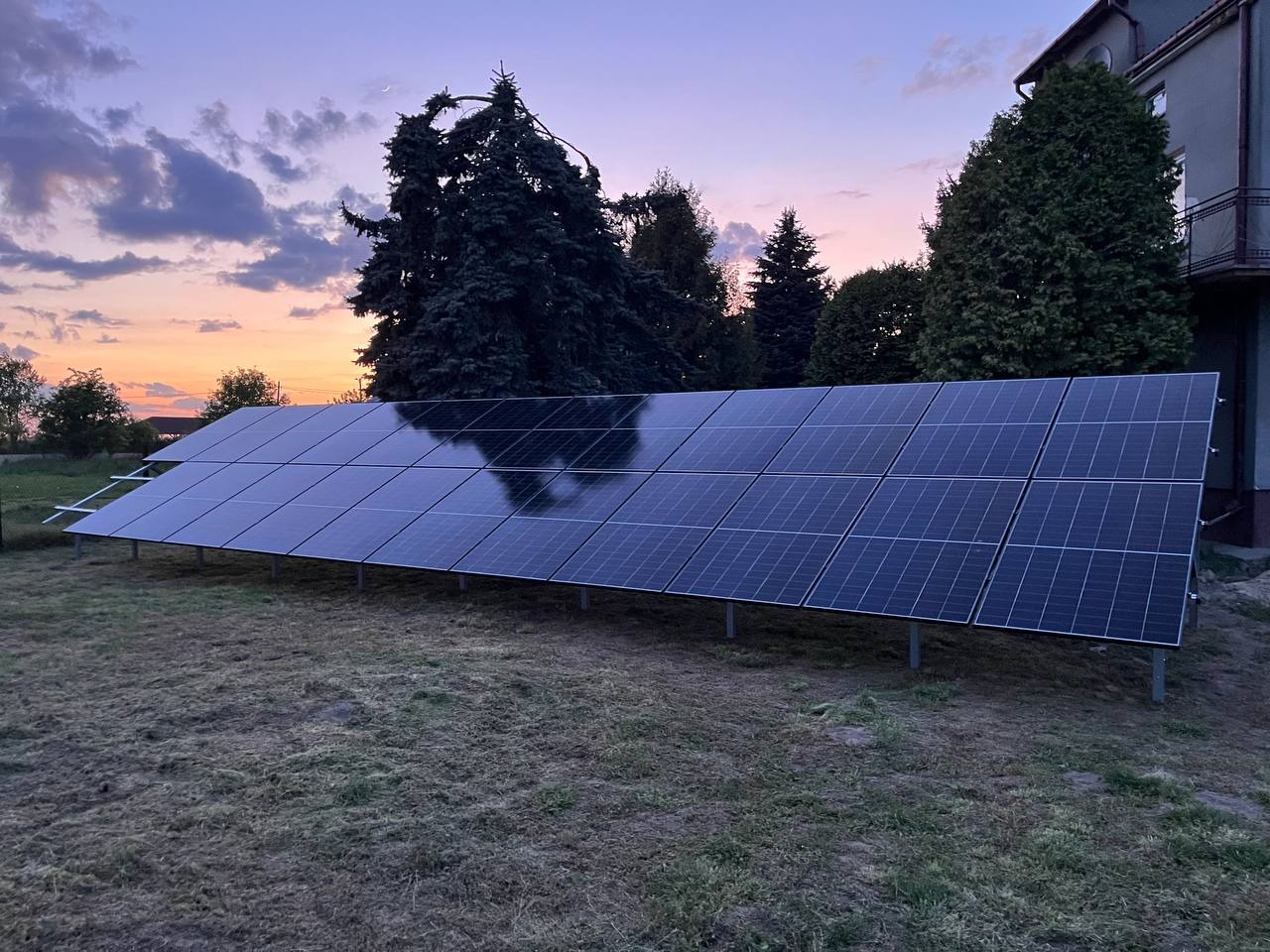 Panel fotowoltaiczny na trawie w Siemianowicach Śląskich, odbijający zachodzące słońce, z budynkiem w tle. Montaż instalacji fotowoltaicznej.
