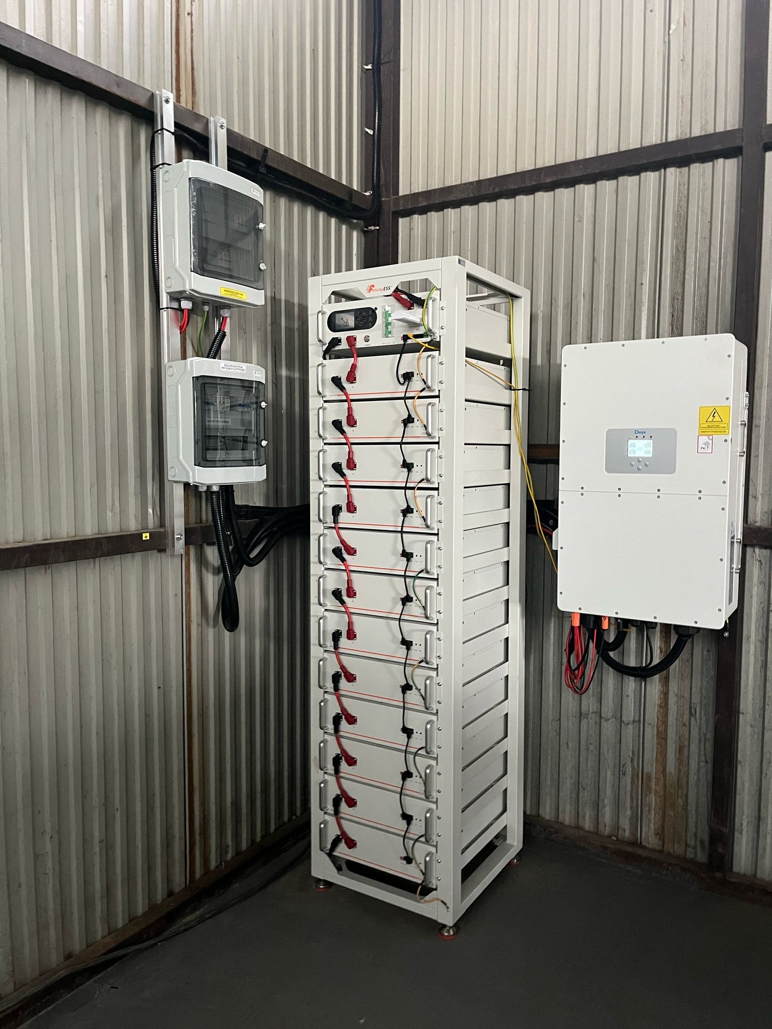 Biała szafa z akumulatorami magazynu energii, skrzynki elektryczne i inwerter na ścianie z blachy falistej w pomieszczeniu. Instalacja gotowa do użytku.