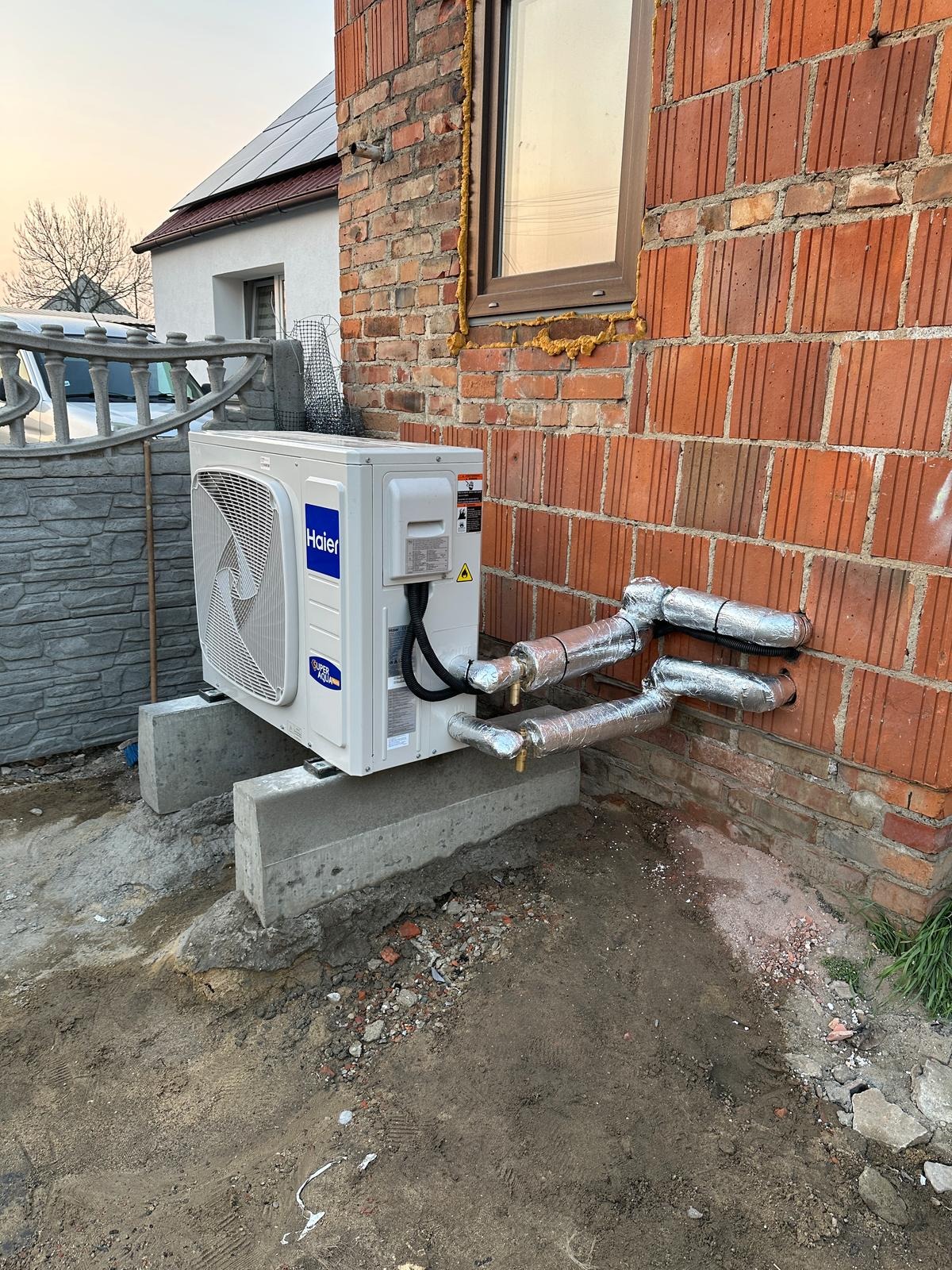 Biała jednostka zewnętrzna klimatyzacji Haier, zamontowana na betonowych podstawach przy ceglanej ścianie budynku. Widoczne ocieplone rury instalacji. Na dachu panele fotowoltaiczne.