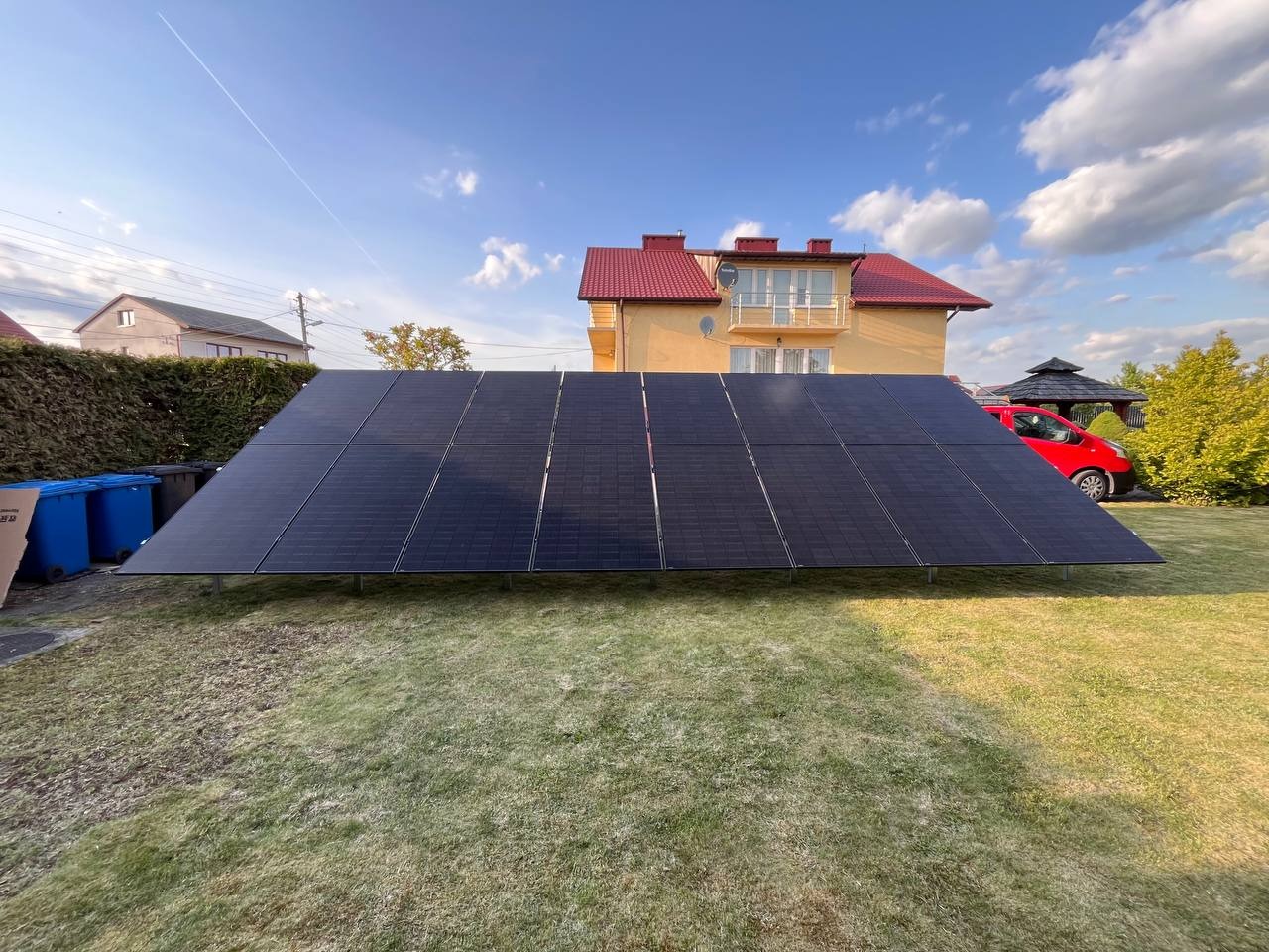 Panele fotowoltaiczne na trawniku przed domem z czerwoną dachówką, obok czerwony samochód i niebieskie pojemniki na śmieci. Błękitne niebo z chmurami.
