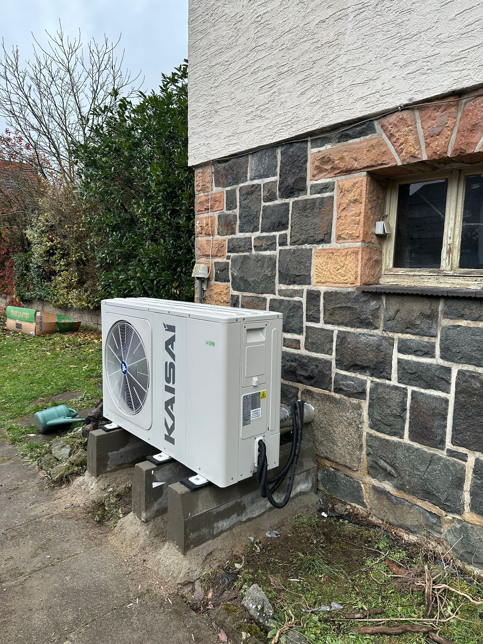 Biała pompa ciepła Kaisai, zamontowana na betonowych wspornikach przy kamiennej ścianie budynku. Widoczne przewody i kartony po urządzeniu w tle.