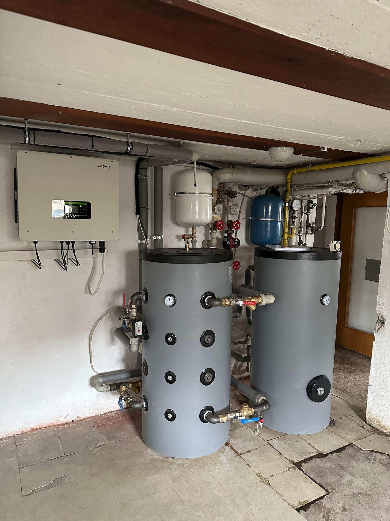 Dwa szare zbiorniki akumulacyjne ciepła z przyłączami, obok inwerter SOFAR. Widoczne rury, instalacja. Całość w pomieszczeniu z płytkami na podłodze.