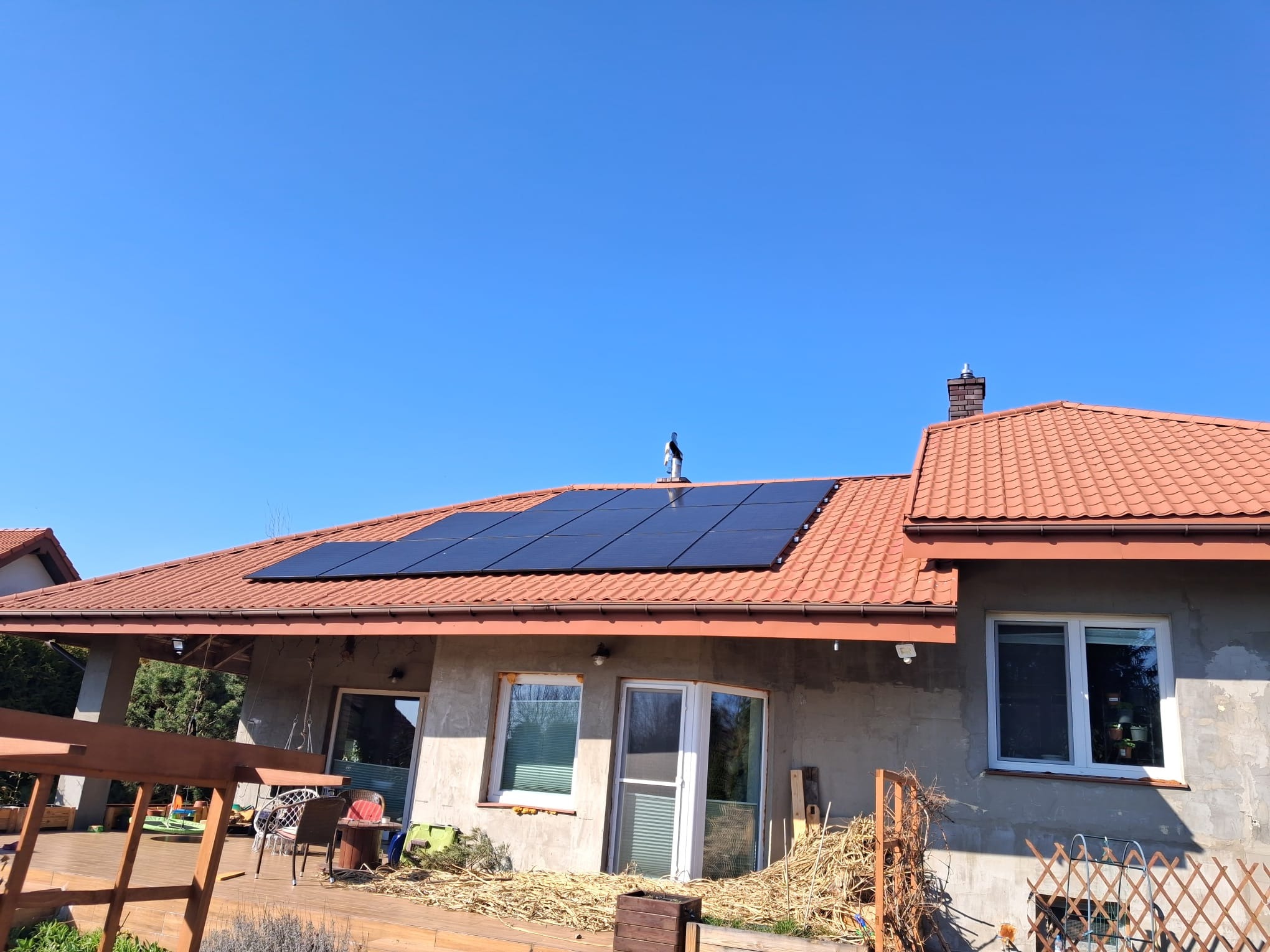 Panel słoneczny na dachu domu z czerwoną dachówką, błękitne niebo. Widok z dołu. Dach lekko skośny. Na kominie siedzi ptak. Częściowo widoczny taras.