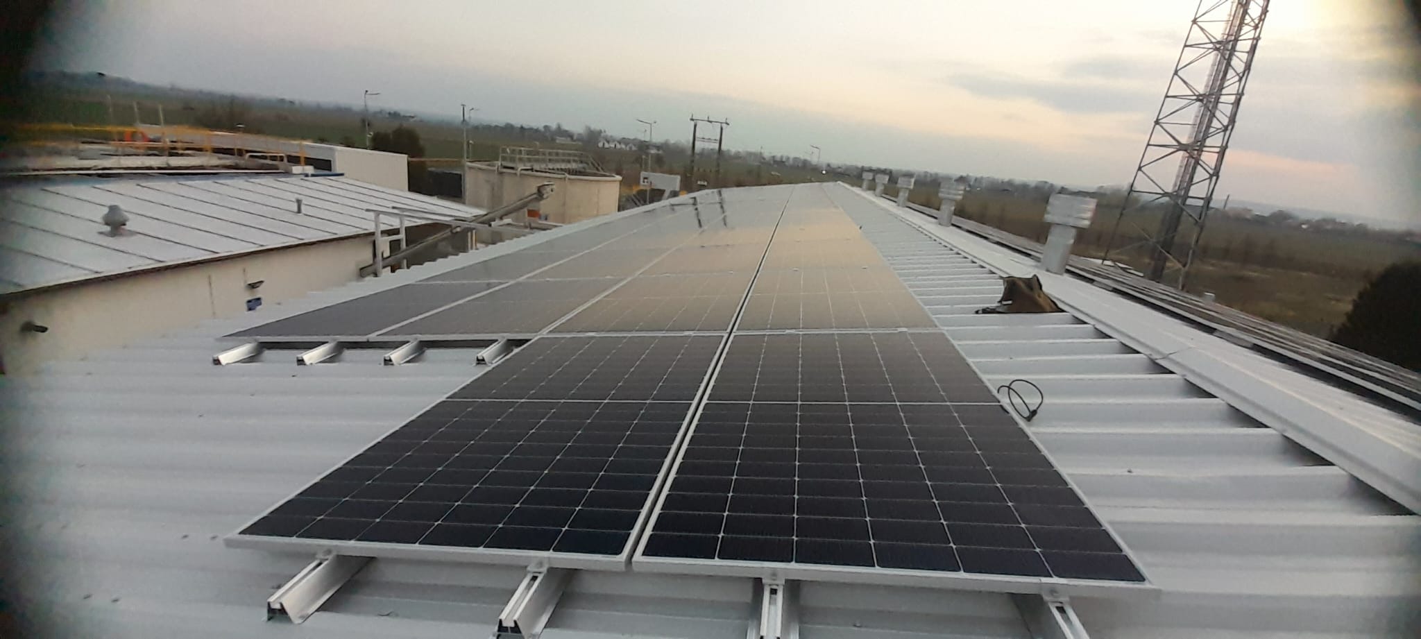 Panele słoneczne na dachu budynku przemysłowego w pochmurny dzień. Widoczna konstrukcja wsporcza paneli i elementy otoczenia.