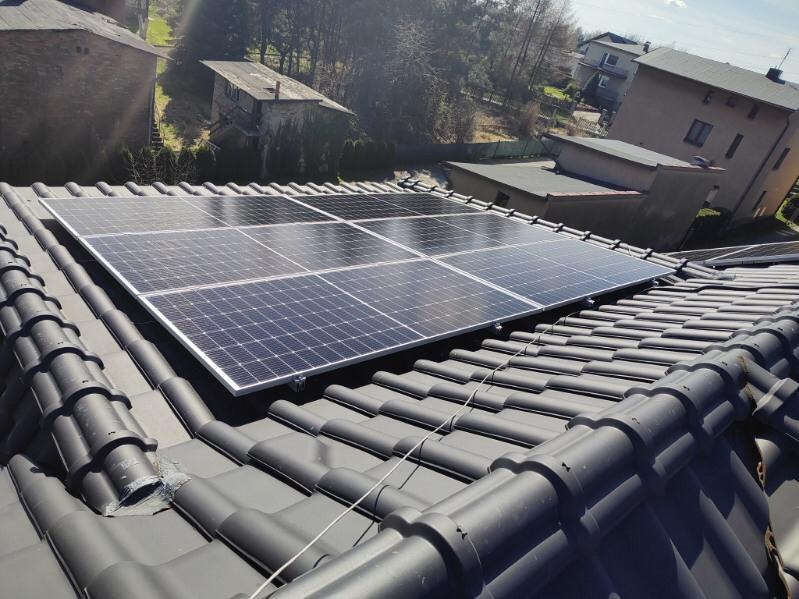 Panel słoneczny na dachu z dachówki ceramicznej w otoczeniu zabudowy mieszkalnej. Widok z góry, oświetlenie dzienne, lekki cień.