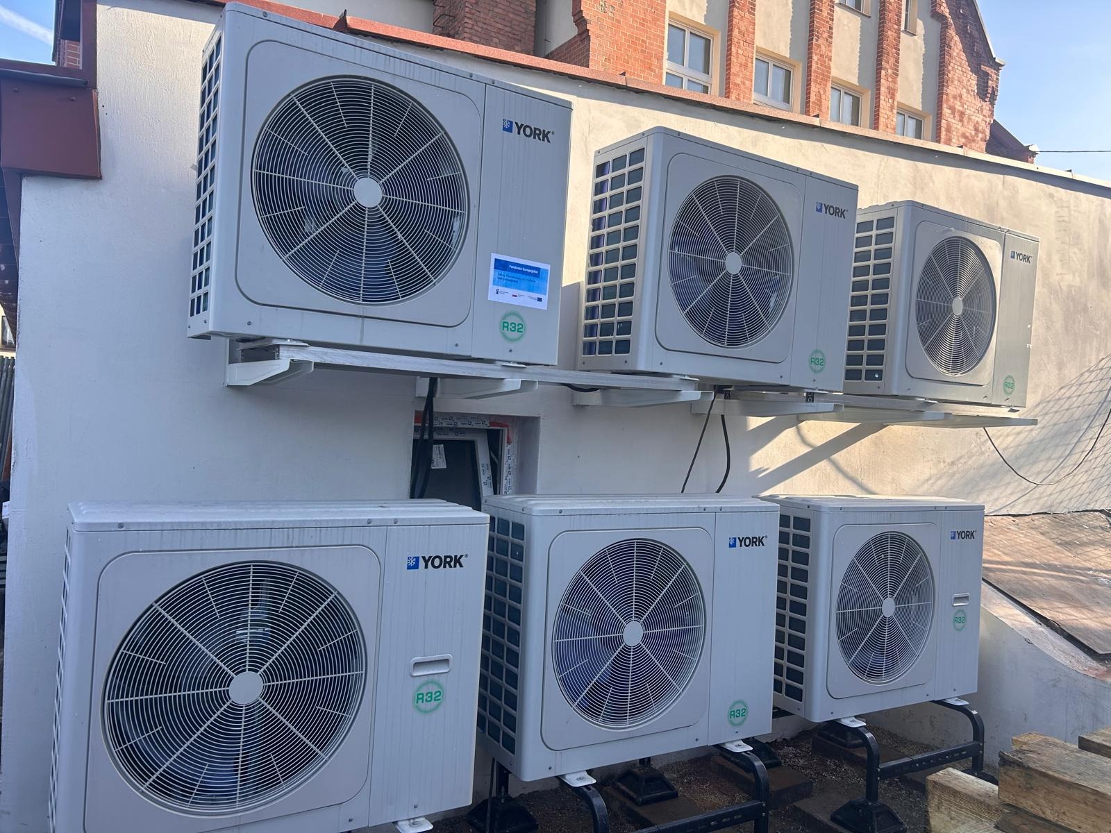 Zewnętrzne jednostki klimatyzacji York zamontowane na ścianie budynku, widoczne metalowe wsporniki i rury. Kilka urządzeń ułożonych w dwóch rzędach, tło stanowi ceglany budynek.