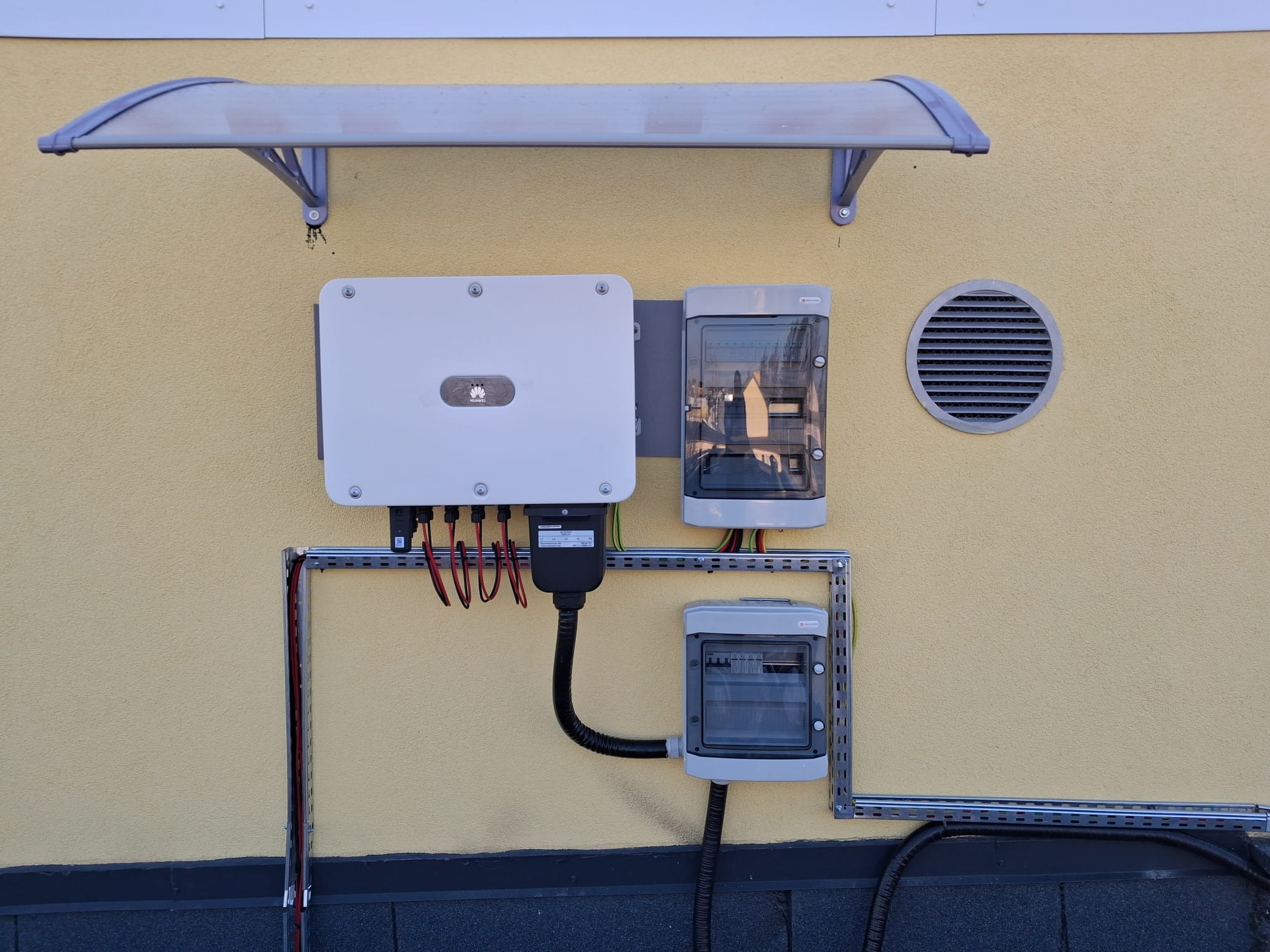 Zamontowany inwerter Huawei z rozdzielnicami na elewacji budynku, widoczne kable i rurki instalacyjne. Nad nimi daszek ochronny i kratka wentylacyjna.