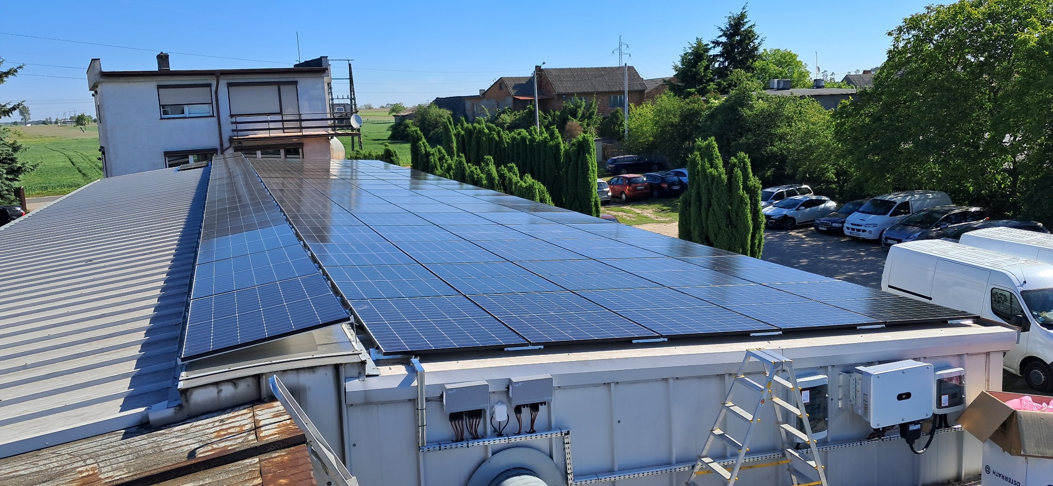 Instalacja fotowoltaiczna na dachu płaskim budynku, widok z góry. Panele ułożone w rzędach, inwertery i skrzynki przyłączeniowe widoczne z boku. Drabina oparta o ścianę.