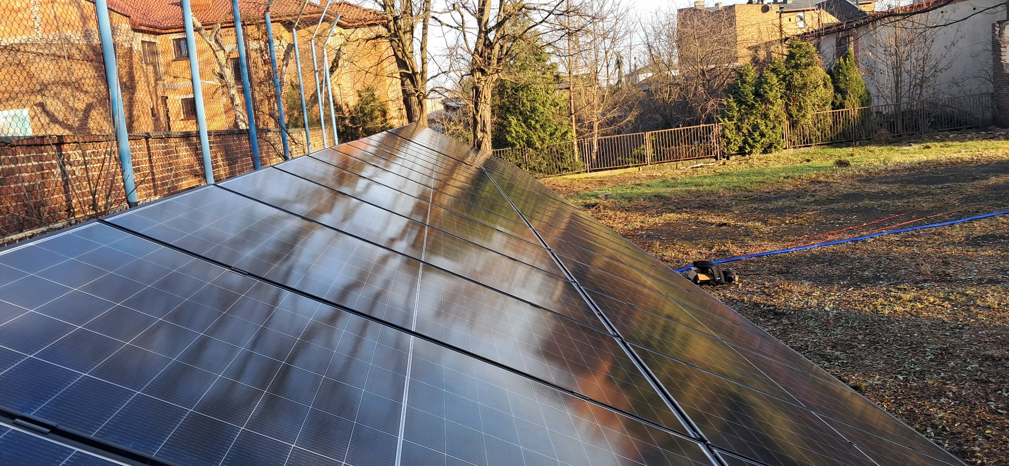 Panel fotowoltaiczny zamontowany na tle budynku i drzew, z widocznymi odblaskami otoczenia na powierzchni paneli. Ujęcie z dołu, podkreślające strukturę instalacji.