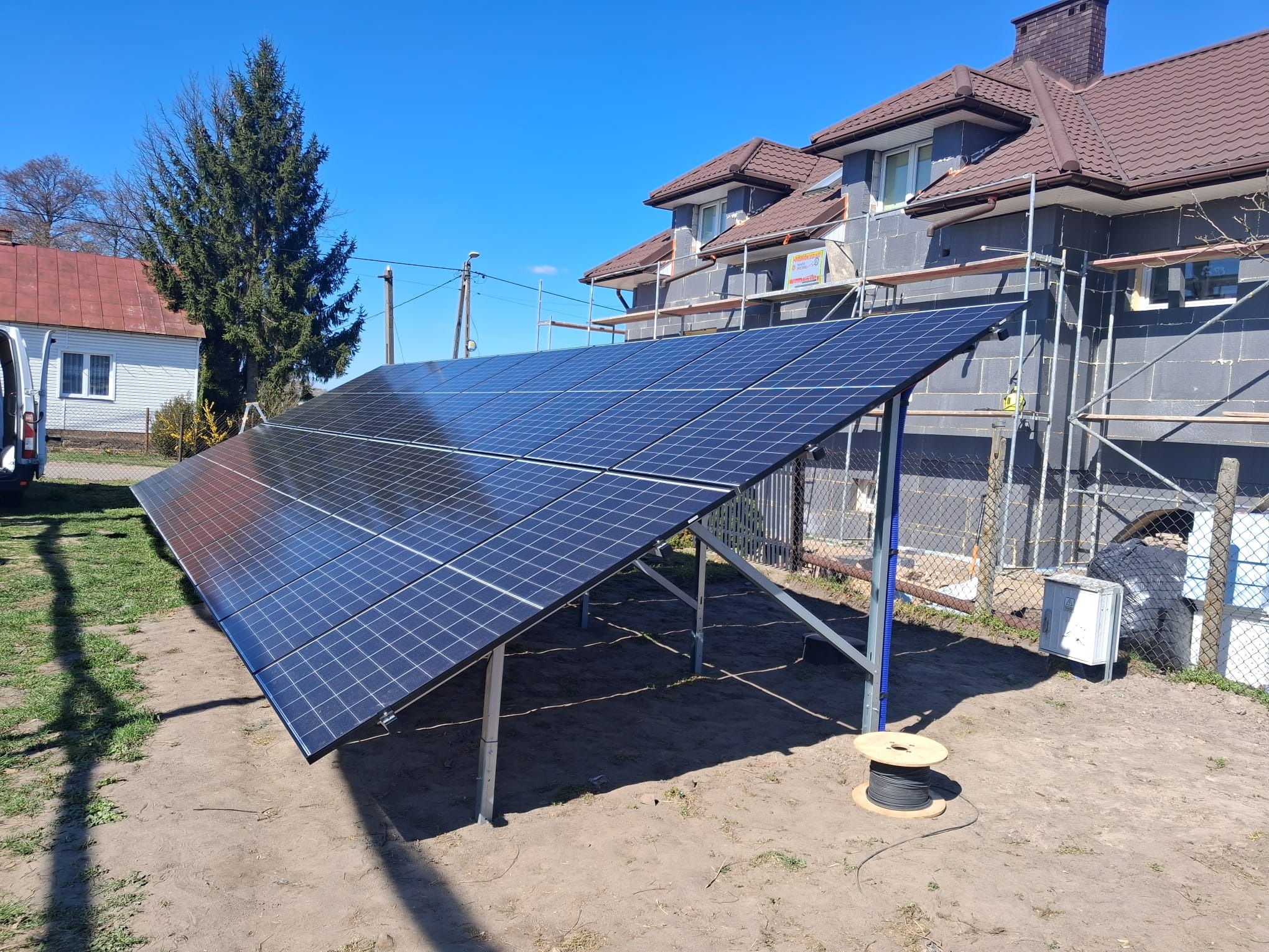 Instalacja paneli słonecznych na konstrukcji wsporczej, widok z boku, dom w trakcie budowy w tle, zwój kabla obok, słoneczny dzień.