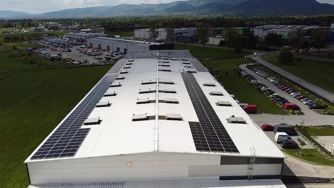 Instalacja fotowoltaiczna na dachu hali magazynowej, widok z góry. Panele wzdłuż krawędzi i w środkowej części dachu. W tle budynki, zieleń i góry.