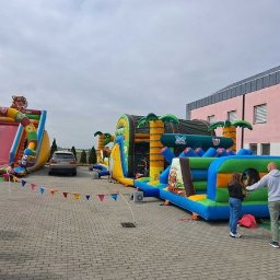 PHU SŁAWOMIR BONK - Kolorowe dmuchańce z motywami dżungli i tygrysa, rozstawione na placu z kostki, z bawiącymi się dziećmi i dekoracyjnymi girlandami. W tle budynek z różową elewacją.