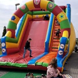 PHU SŁAWOMIR BONK - Kolorowa dmuchana zjeżdżalnia z motywem kwiatów, na której bawią się dzieci. Obok siedzi kobieta na czerwonym stołku. Na zielonym dywanie leżą buty.