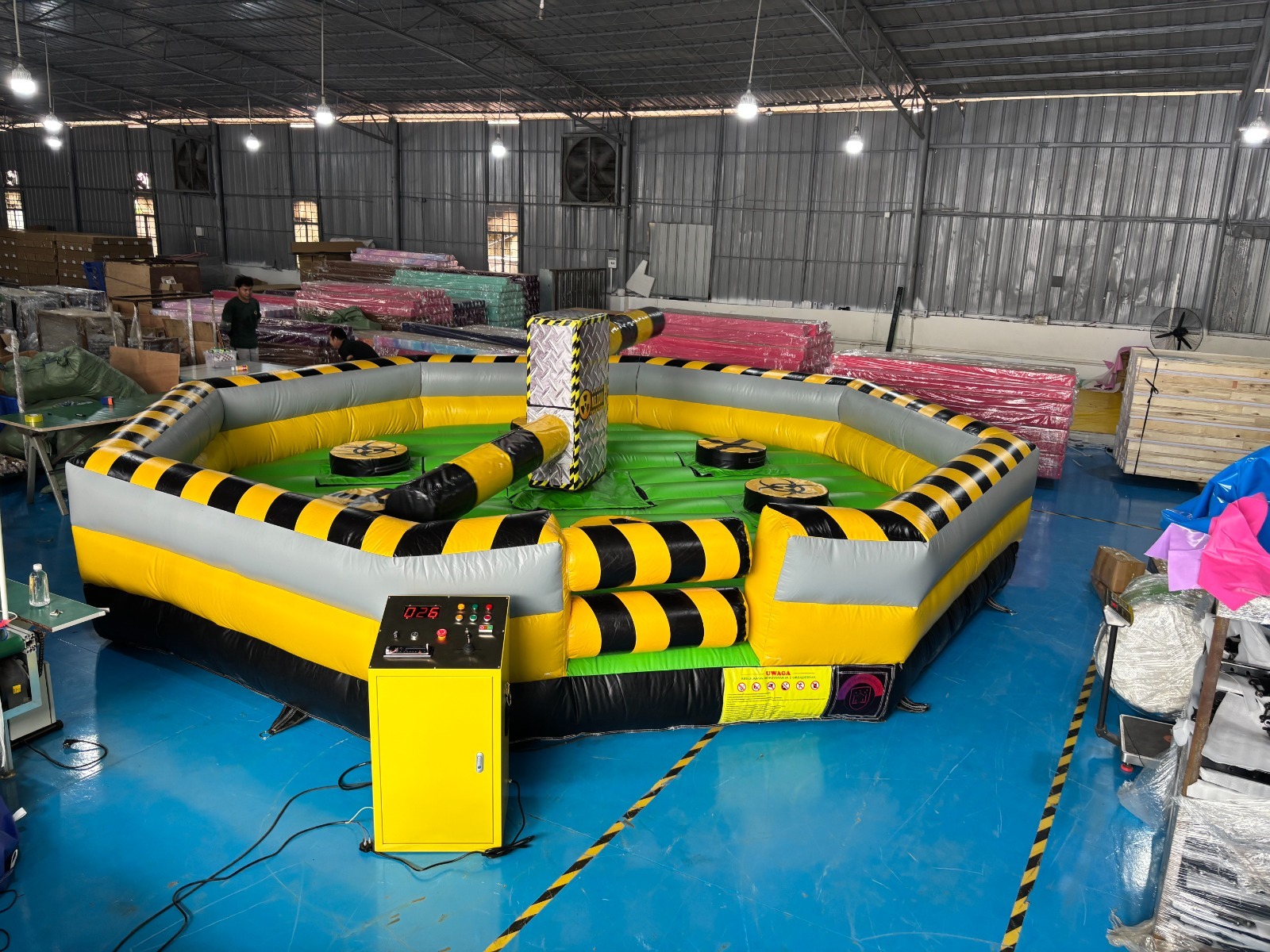 Duży, nadmuchiwany, okrągły tor przeszkód w kolorach żółtym, szarym i czarnym, z centralną konstrukcją i kontrolną konsolą, ustawiony w hali.