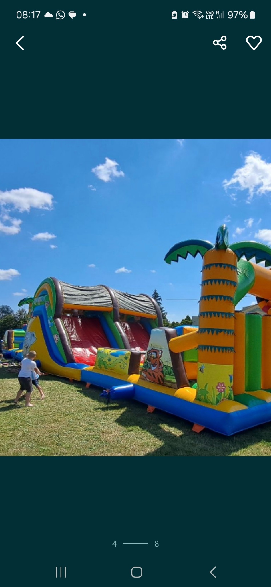 Długi, kolorowy dmuchany tor przeszkód z motywami dżungli na trawie, zjeżdżalnia i dekoracje w kształcie palm. Dzieci w pobliżu. Błękitne niebo w tle.