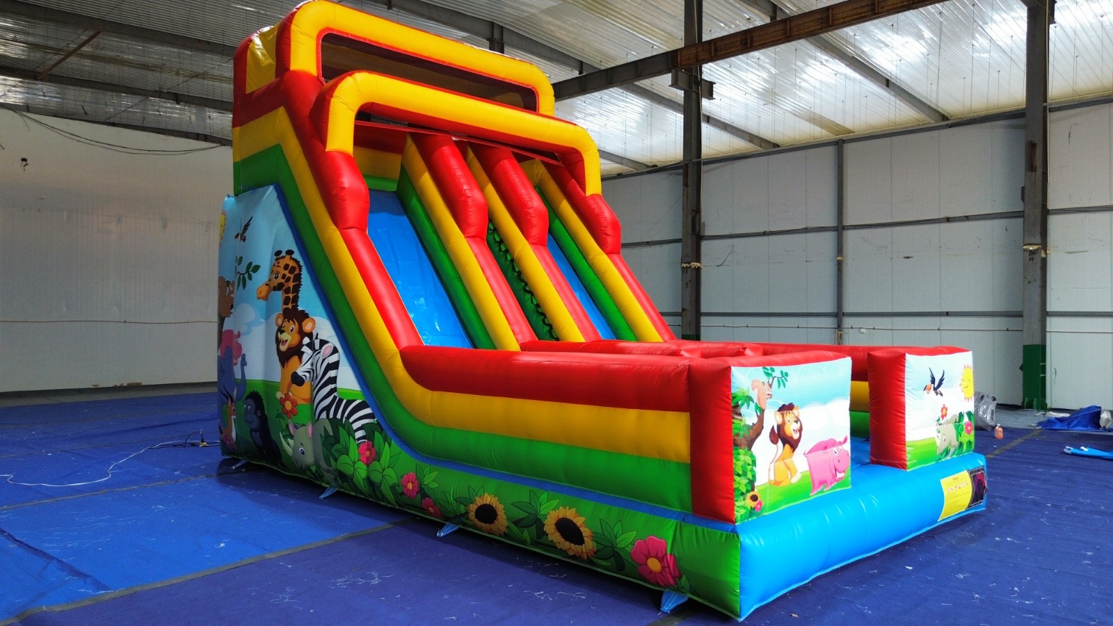 Kolorowa, dmuchana zjeżdżalnia z motywem zwierząt w Gnieźnie. Atrakcja dla dzieci na imprezy plenerowe, widok z boku, w hali z metalową konstrukcją.