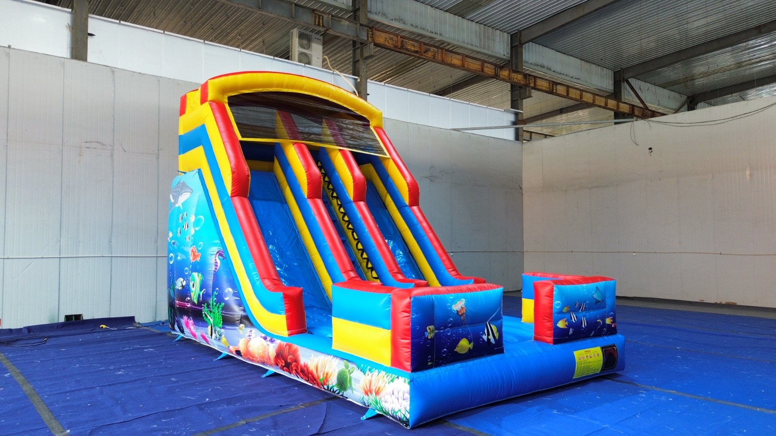 Kolorowa, dmuchana zjeżdżalnia z motywem podwodnego świata, ustawiona na niebieskiej płachcie w hali. Zjeżdżalnia ma trzy tory i jest gotowa do zabawy.
