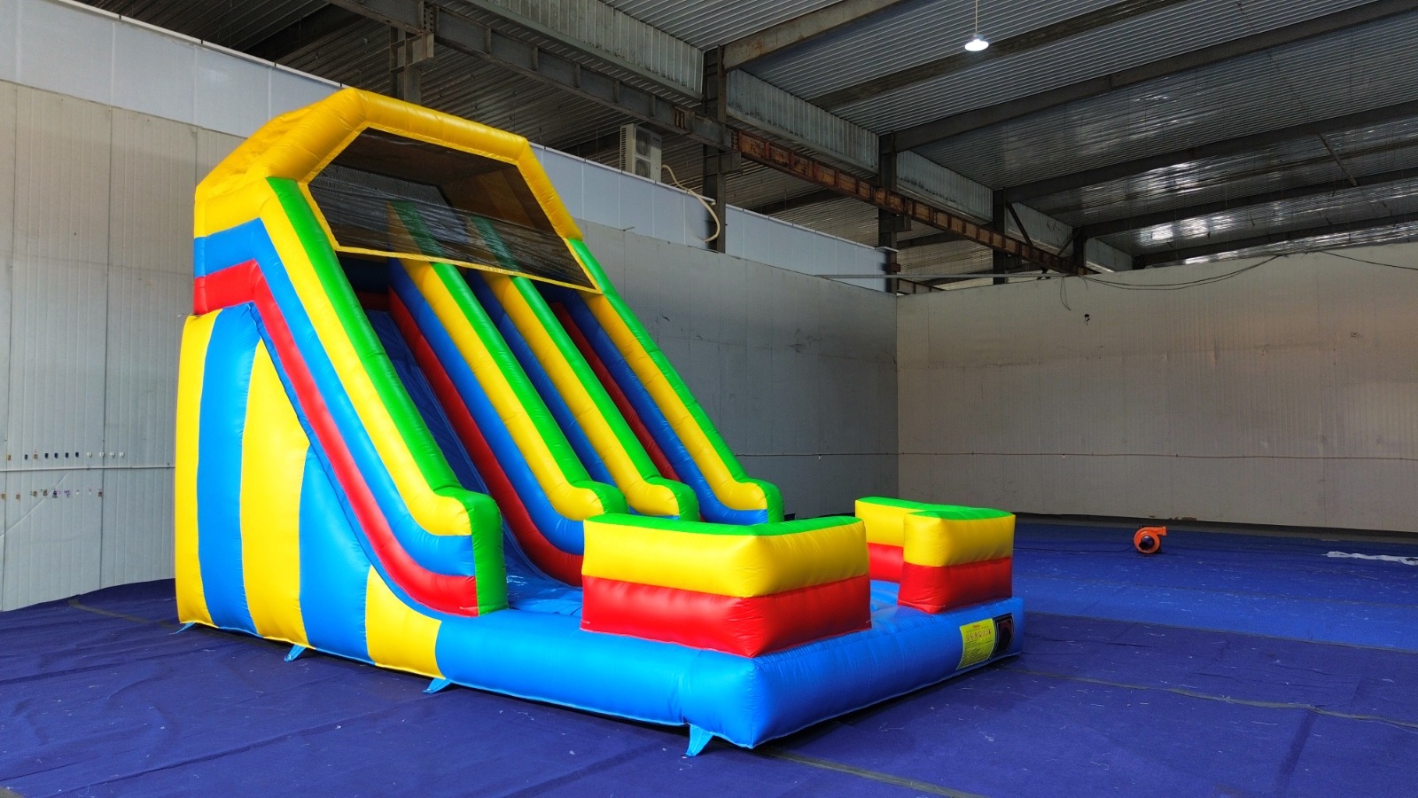 Kolorowa, nadmuchiwana zjeżdżalnia dla dzieci w hali. Widoczny wentylator. Zjeżdżalnia w odcieniach żółci, czerwieni, zieleni i niebieskiego. Podłoże pokryte niebieską plandeką.