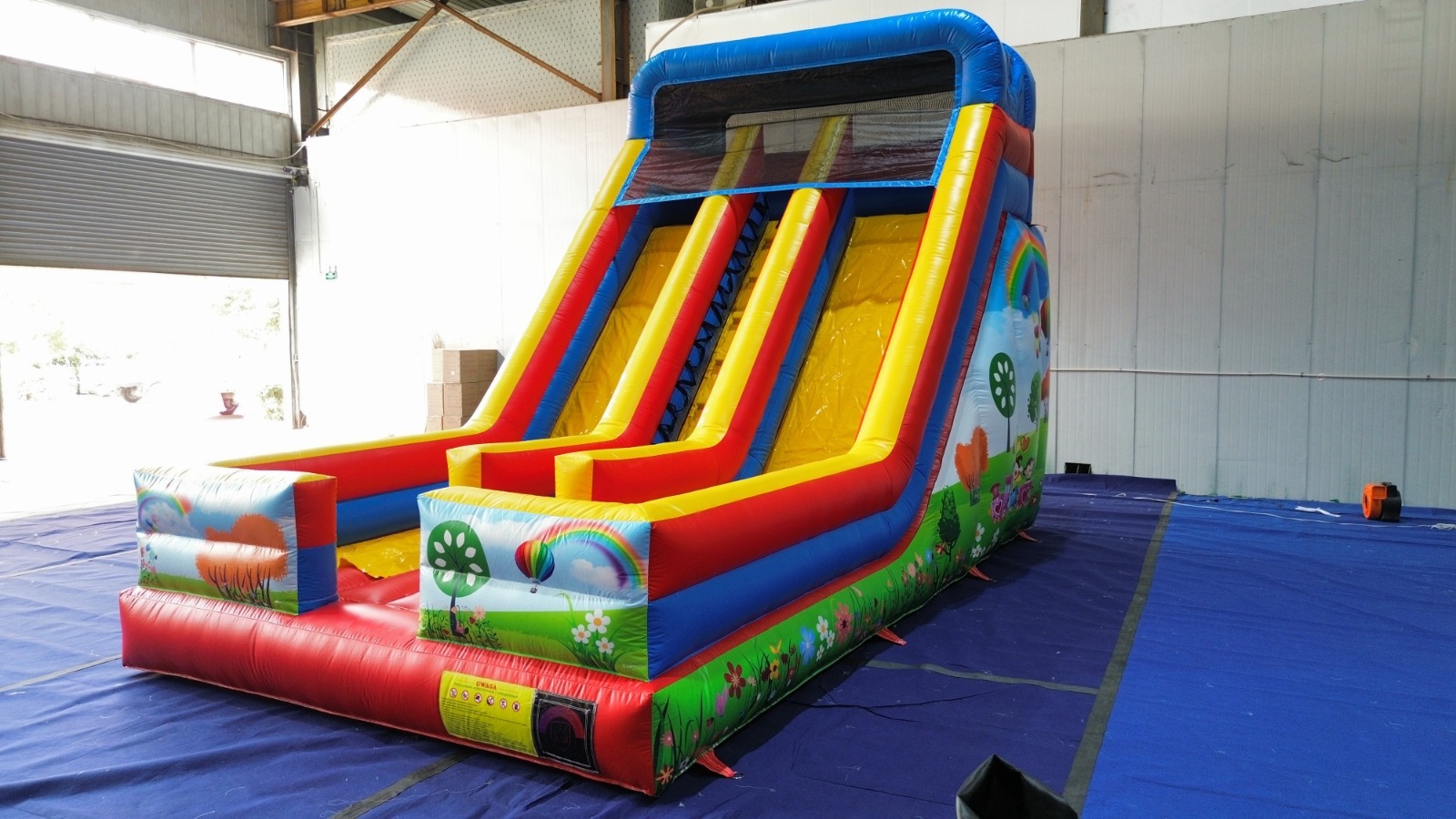 Kolorowa, dmuchana zjeżdżalnia z dwoma torami, z motywami roślinnymi i tęczy, ustawiona na niebieskiej płachcie w hali z blach falistych w tle.