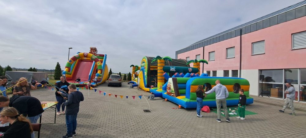 Kolorowe dmuchańce z motywami dżungli i tygrysa, rozstawione na placu z kostki, z bawiącymi się dziećmi i dekoracyjnymi girlandami. W tle budynek z różową elewacją.