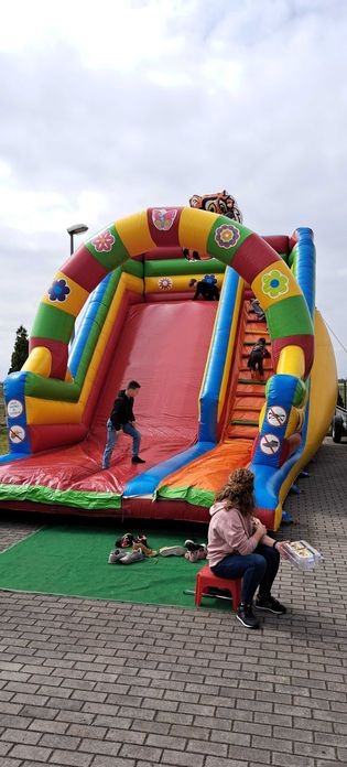 Kolorowa dmuchana zjeżdżalnia z motywem kwiatów, na której bawią się dzieci. Obok siedzi kobieta na czerwonym stołku. Na zielonym dywanie leżą buty.