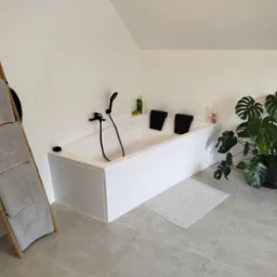 Minimalistyczna łazienka na poddaszu: biała wanna z czarną armaturą, drabina z ręcznikami i duża monstera. Jasne płytki podłogowe.