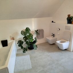 Wykończenia wnętrz - Jasna łazienka na poddaszu: biała wanna z czarnymi zagłówkami, bidet i toaleta w zabudowie. Akcenty roślinne: monstera i rośliny na półce. Szare płytki podłogowe.