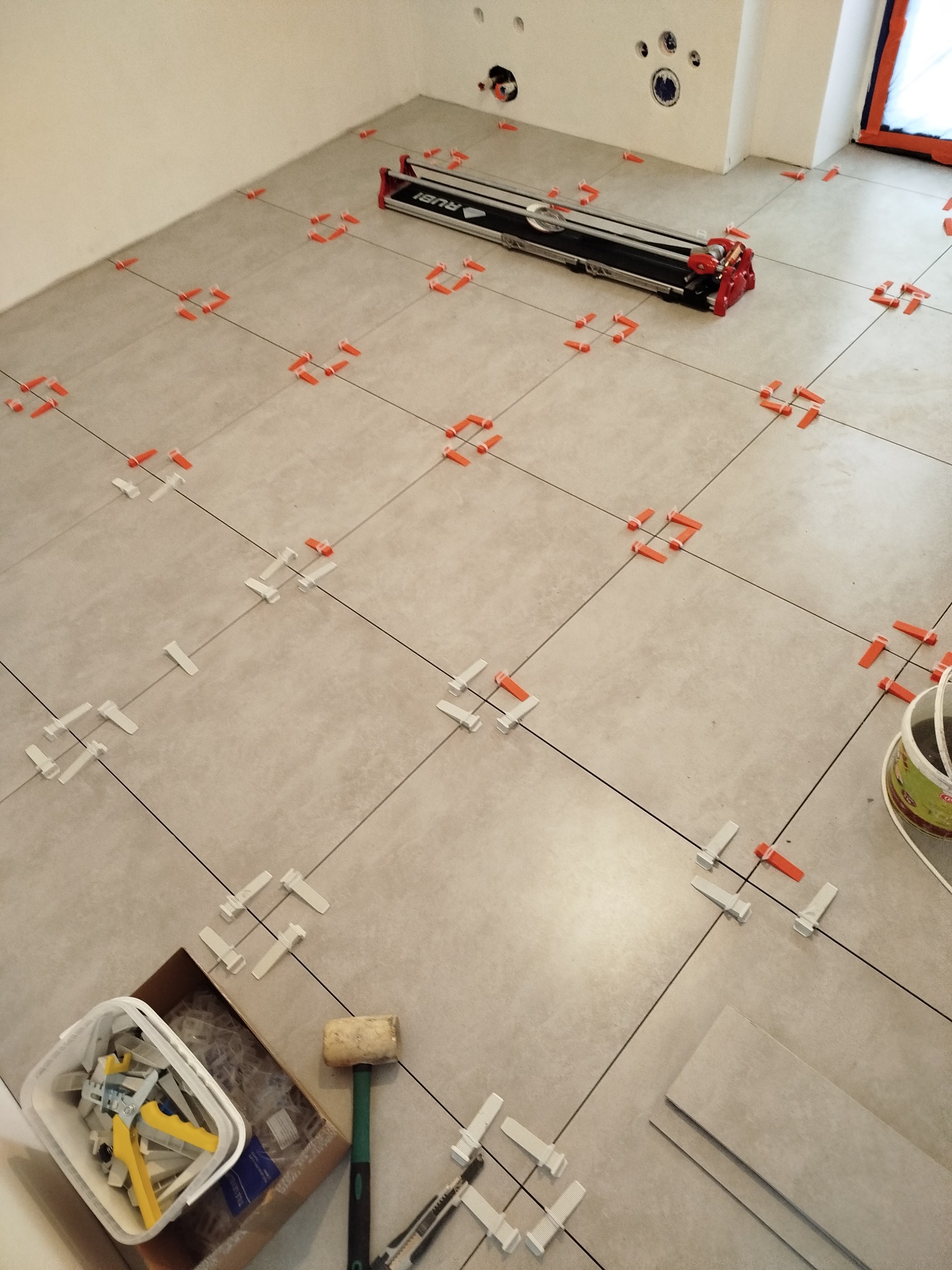 Układanie jasnych, kwadratowych płytek podłogowych z użyciem systemu poziomowania. Widoczna maszynka do cięcia płytek i narzędzia glazurnicze.