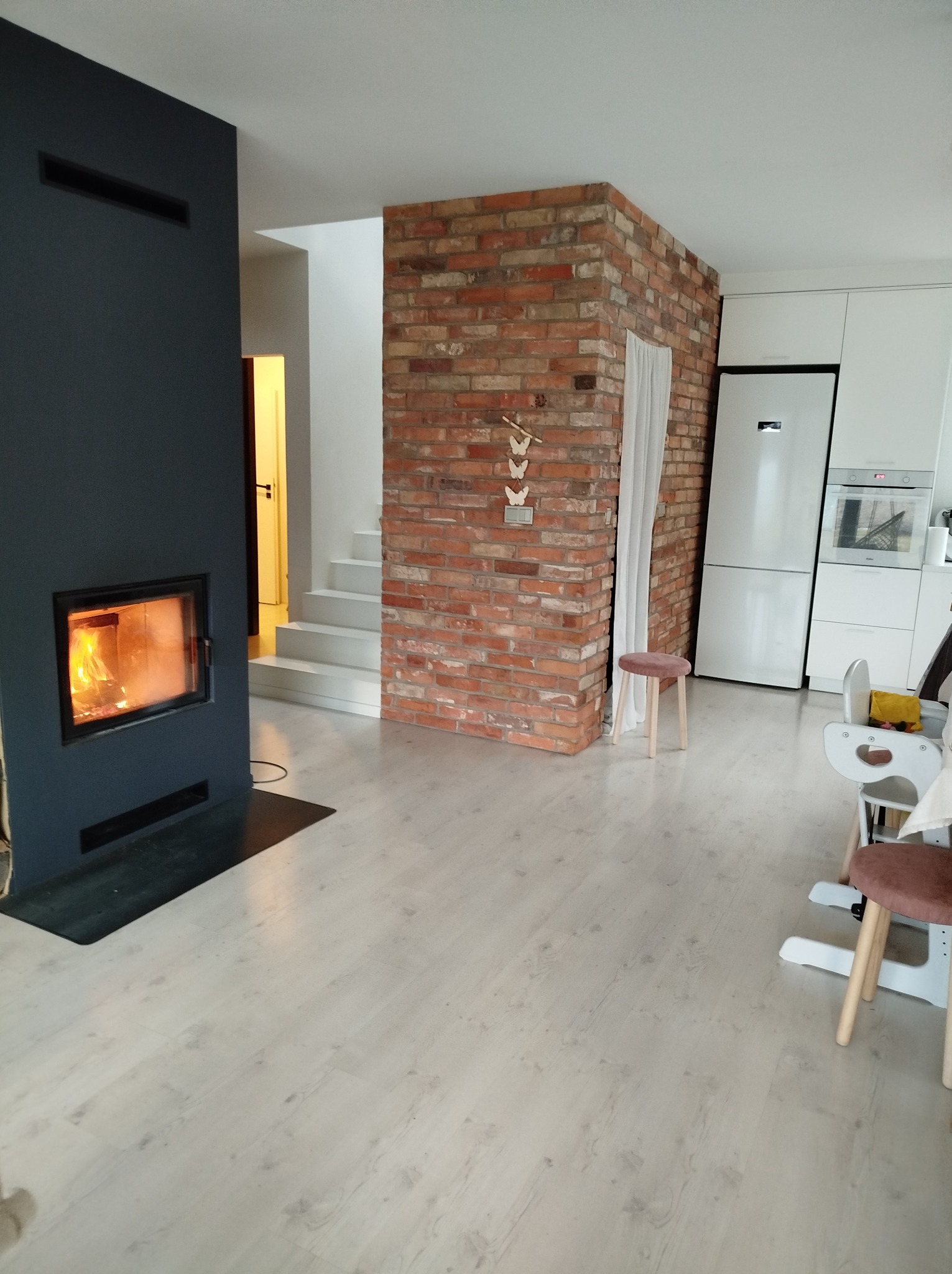 Salon z kominkiem w nowoczesnym stylu, ściana z cegły, jasna podłoga i schody. Widoczne elementy wyposażenia kuchni i krzesełka dziecięce. Ciepłe światło z kominka.