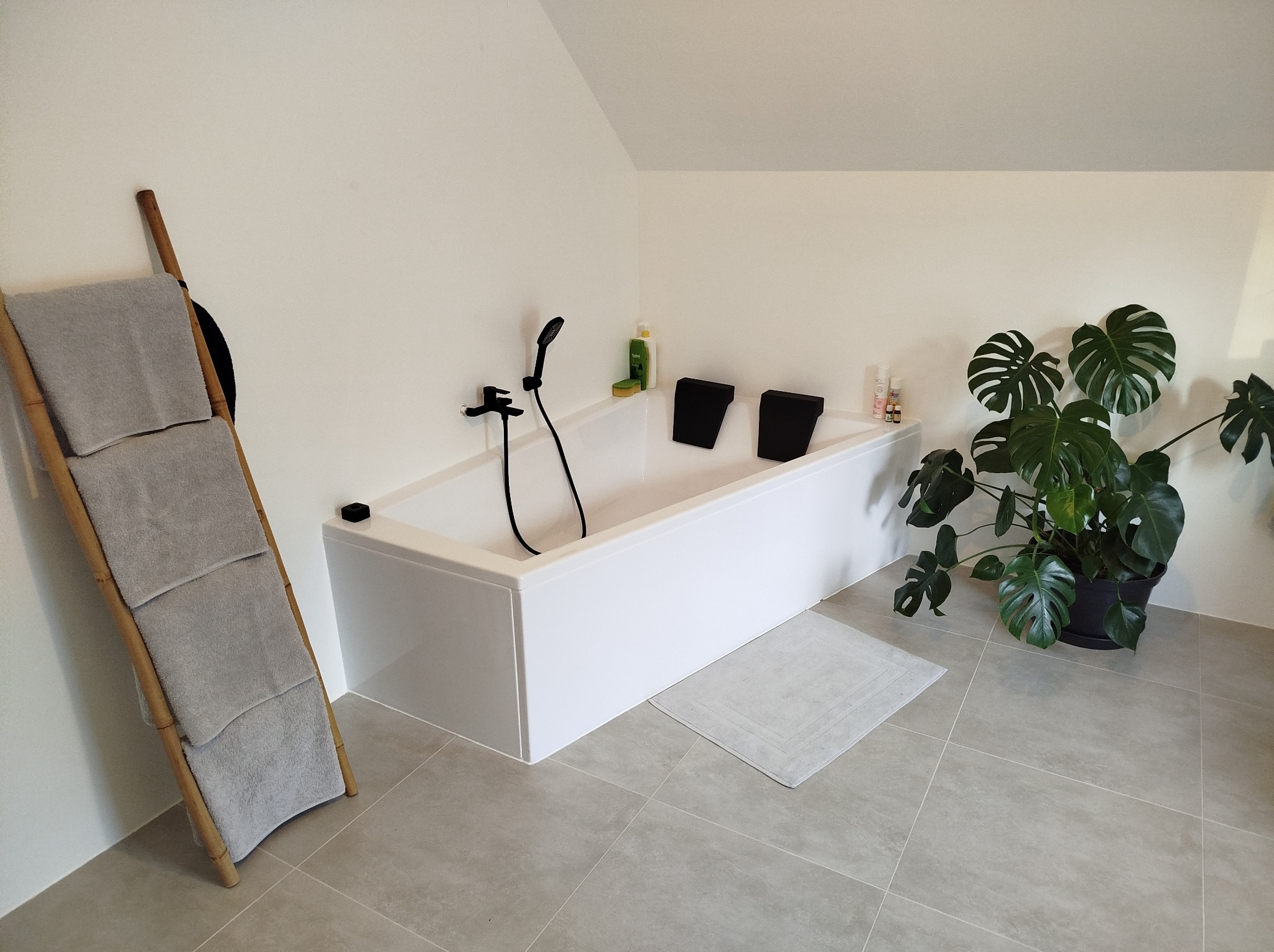 Minimalistyczna łazienka na poddaszu: biała wanna z czarną armaturą, drabina z ręcznikami i duża monstera. Jasne płytki podłogowe.