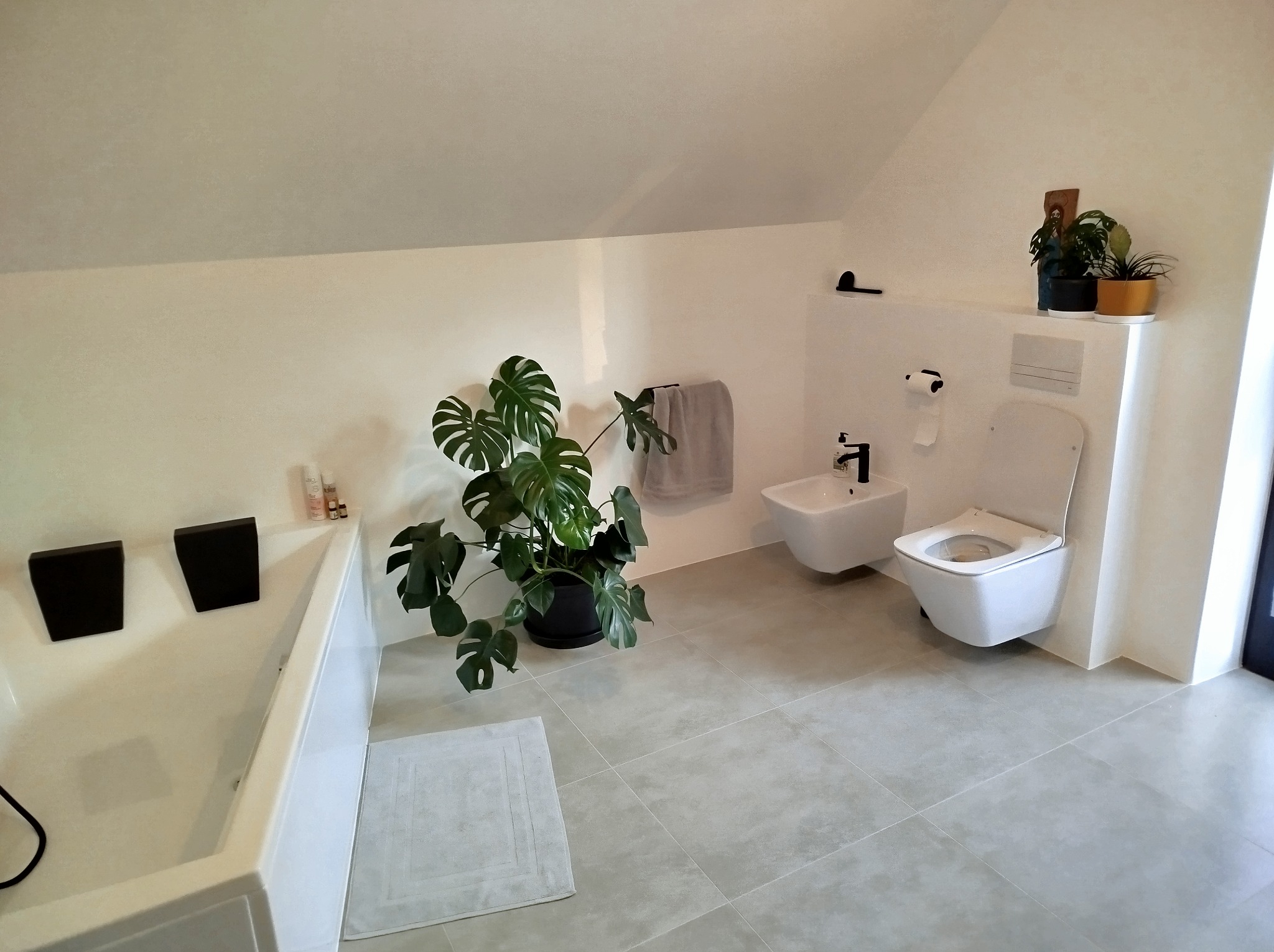 Jasna łazienka na poddaszu: biała wanna z czarnymi zagłówkami, bidet i toaleta w zabudowie. Akcenty roślinne: monstera i rośliny na półce. Szare płytki podłogowe.