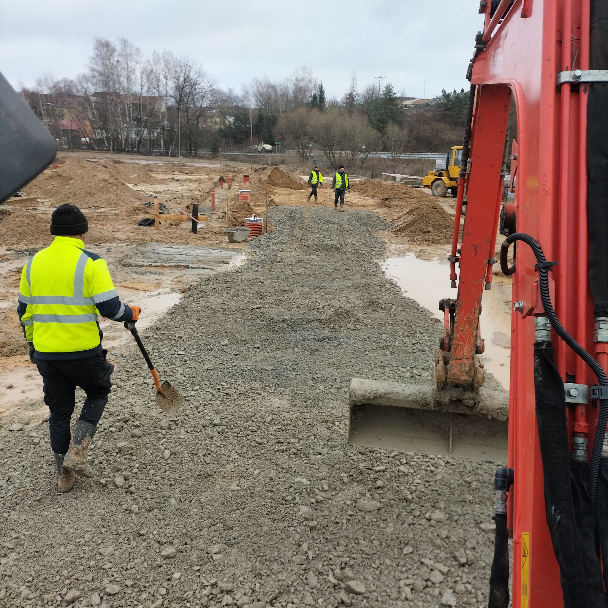 Prace ziemne w toku: robotnicy w odblaskowych kamizelkach, koparka i przygotowane podłoże z gruzu na terenie budowy w pochmurny dzień.