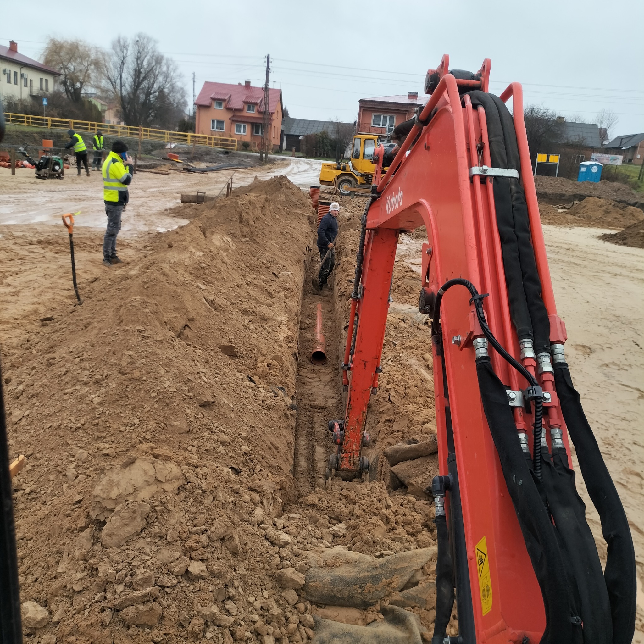 Pomarańczowa koparka Kubota w wykopie ziemnym, robotnicy układają rury. W tle zabudowa wiejska i sprzęt budowlany. Prace ziemne w toku.