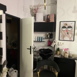 Serhii Burlin - Nowoczesne wnętrze salonu fryzjerskiego z czarnym fotelem, umywalką, półkami z kosmetykami i plakatem z wizerunkiem kobiety w okularach. Widoczne otwarte drzwi.