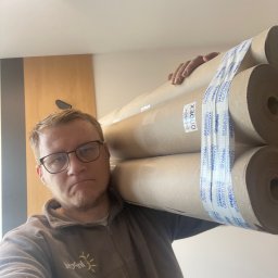 Serhii Burlin - Mężczyzna w okularach niesie na ramieniu kilka rolek materiału budowlanego, prawdopodobnie do wykończenia wnętrz. Widoczna etykieta 'DODEX'.