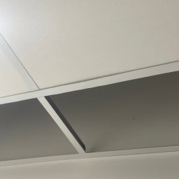 Serhii Burlin - Zamontowany sufit podwieszany z kasetonami, widok z dołu. Białe profile i szare panele tworzą geometryczny wzór. Widoczne niedoskonałości na panelu.