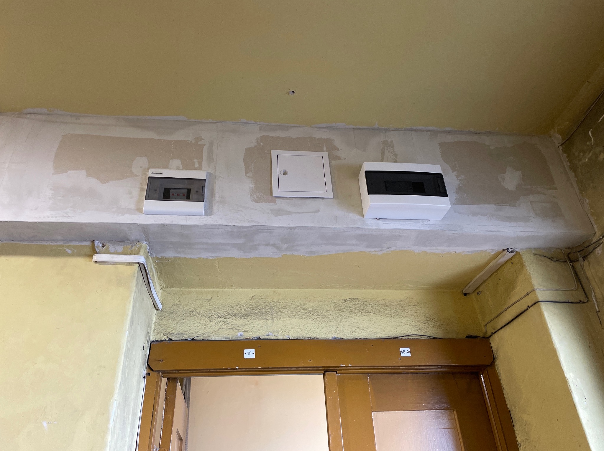 Instalacja elektryczna: trzy białe skrzynki na ścianie, pośrodku mała kwadratowa. Ściana w trakcie prac wykończeniowych, widoczne ślady po szpachlowaniu. Nad drzwiami.