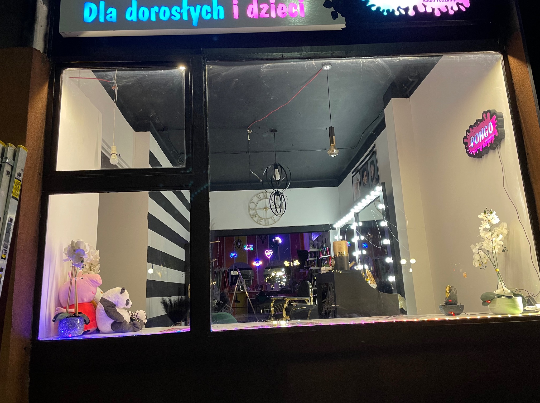 Widok przez okno salonu z neonami i dekoracjami, widoczne zabawki i akcesoria, oświetlenie dekoracyjne.