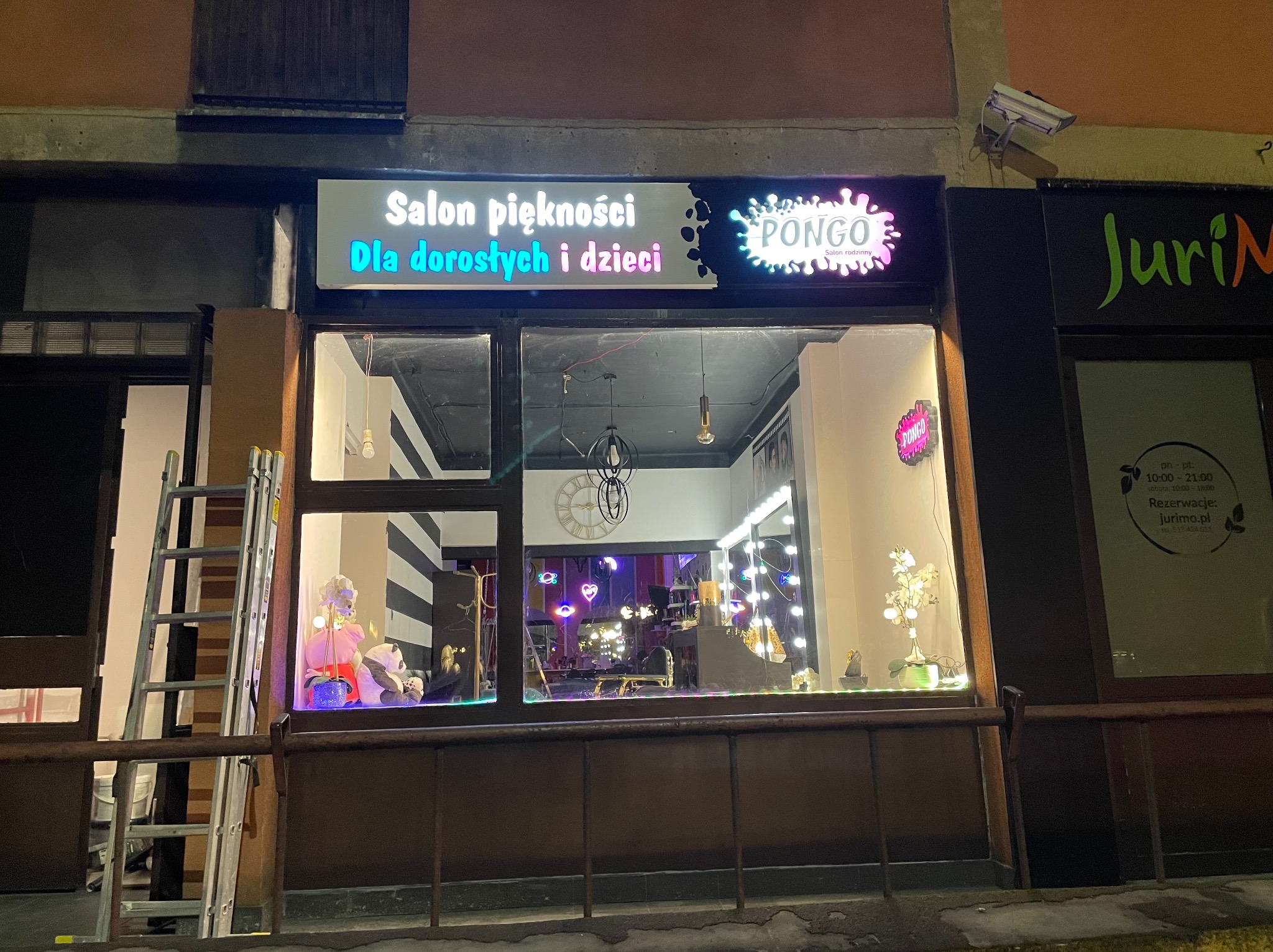 Fasada salonu piękności 'Pongo' z oknem wystawowym, widoczny szyld z napisem 'Salon piękności dla dorosłych i dzieci', dekoracje świetlne i schody przystawione do wejścia.